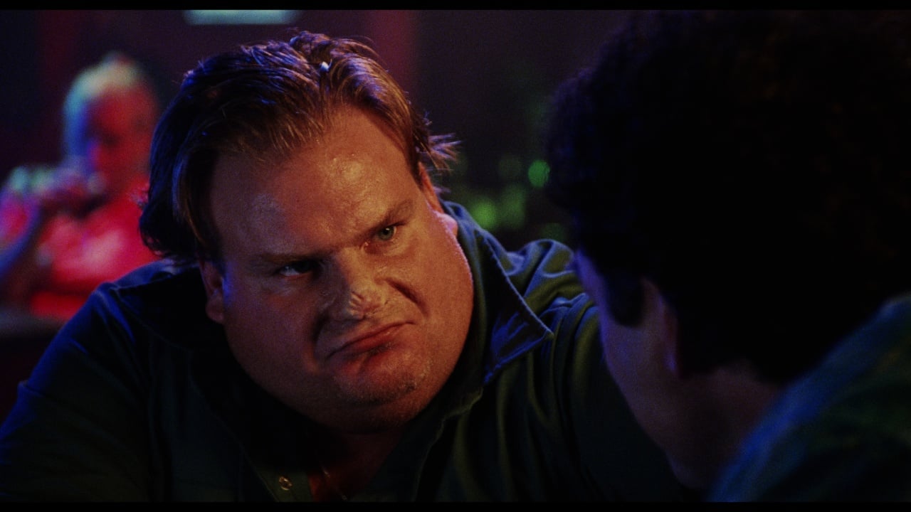 A man with a stern expression sits at a table in a dimly lit setting, facing another person whose back is to the camera.