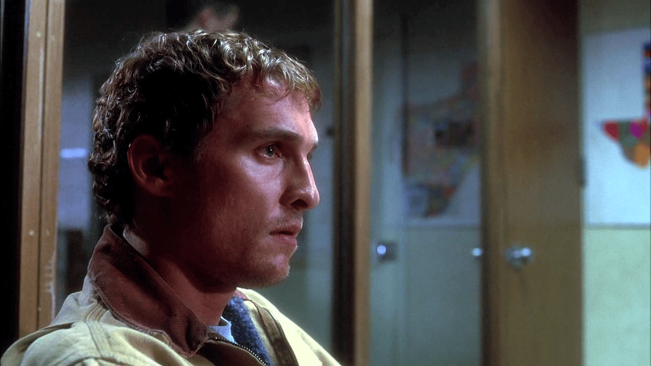 A man with short, curly hair and a serious expression sits indoors, facing left, with blurred office partitions and wall decorations in the background.