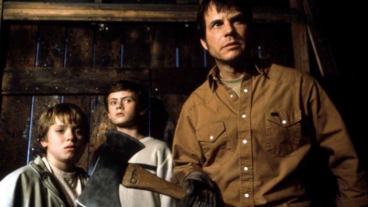 A man holding an axe stands in front of two boys, all looking in the same direction, inside a dimly lit wooden structure.