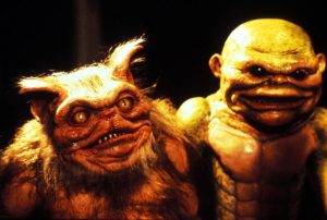 Two detailed animatronic monster puppets stand side by side; one is furry with horns and sharp teeth, the other is green, hairless, and has scaly skin and a broad mouth.