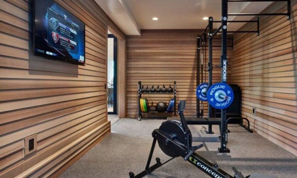 Home gym room with wooden walls, an exercise bike, weightlifting rack, dumbbells, weight plates, and a wall-mounted TV displaying a sports game.