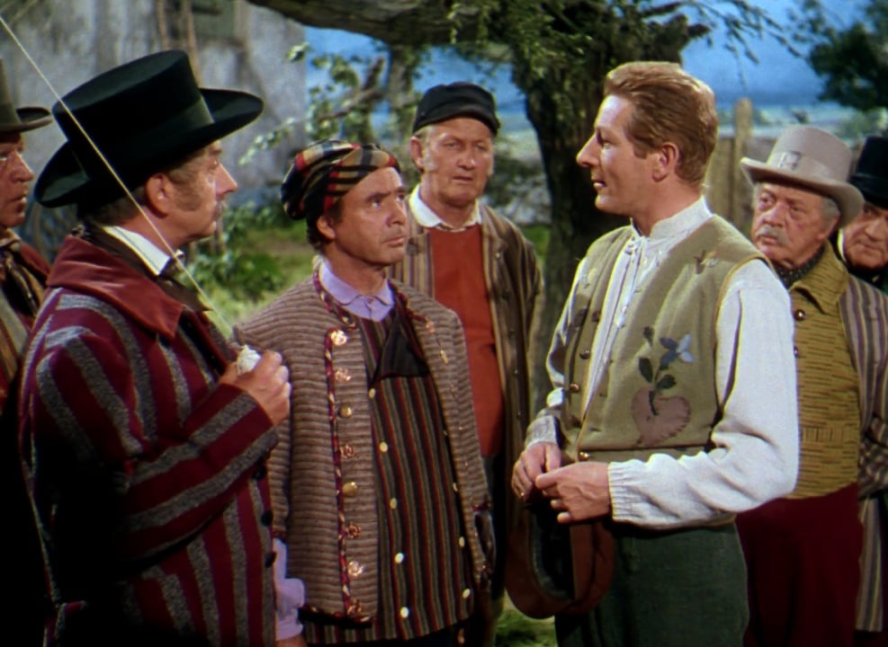 A group of men in vintage, colorful clothing stand outdoors, engaged in conversation; one man holds a hat, and another holds a fencing foil.