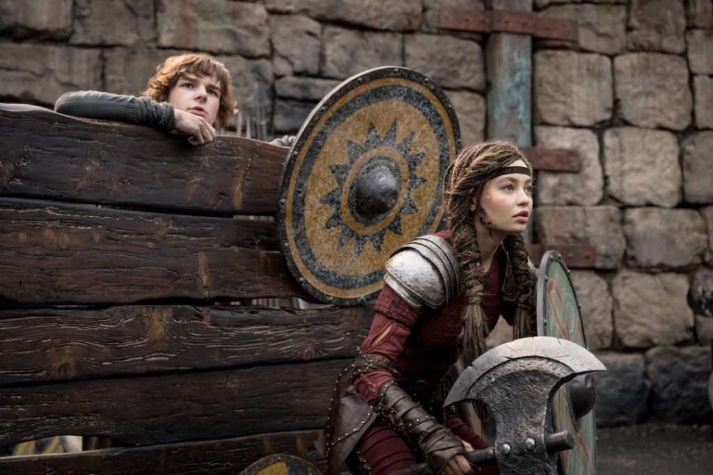 Two young people in medieval-style armor and clothing hold shields and weapons beside a wooden barrier, with a stone wall in the background.
