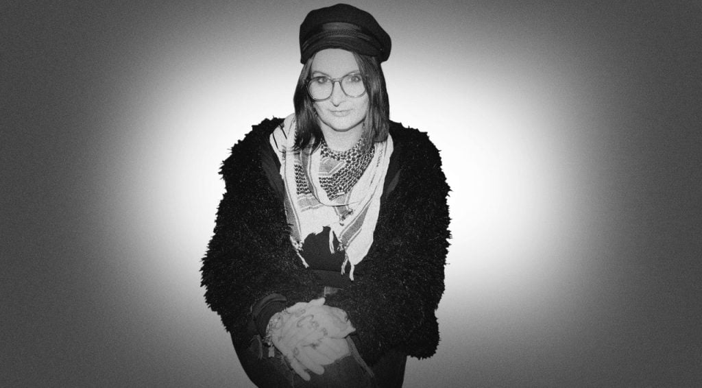 Person wearing glasses, a cap, a patterned scarf, and a textured jacket sits with hands clasped against a plain, gradient background.