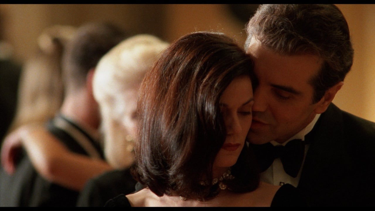 A man in a tuxedo leans in close to a woman in a black dress at a formal event, with another couple dancing in the blurred background.