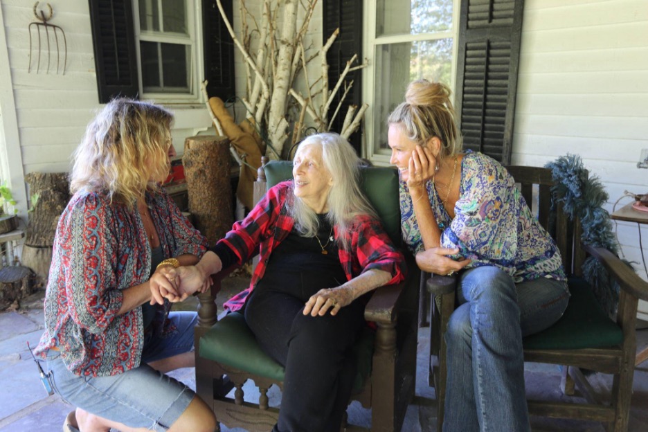 Three women sit and talk on a porch, with one elderly woman in the center holding hands and smiling at the two younger women beside her.