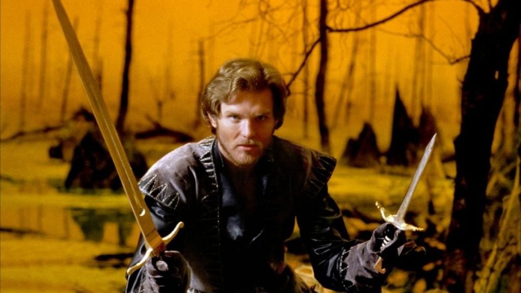 A man in medieval-style armor holds two swords in a defensive stance in a swamp with bare trees and an orange sky.