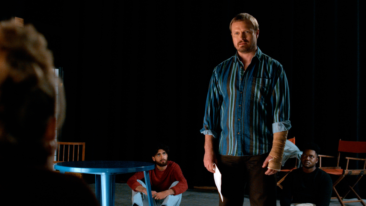 A man stands holding papers, facing a seated person, while two others sit on the floor behind him in a dimly lit room with black curtains and scattered chairs.