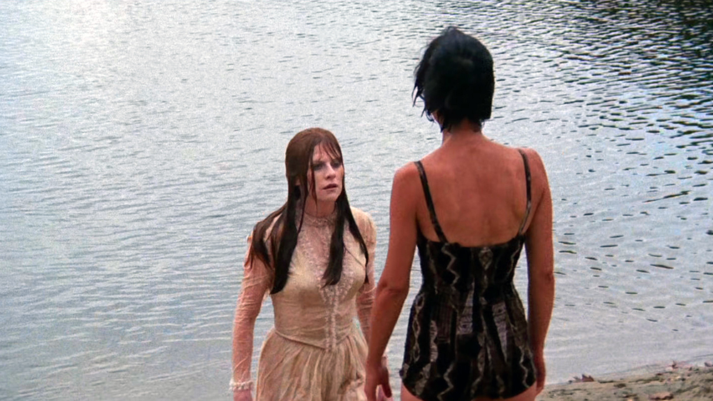 A woman in a wet, long white dress stands in shallow water facing another woman in a black swimsuit on a sandy shore.
