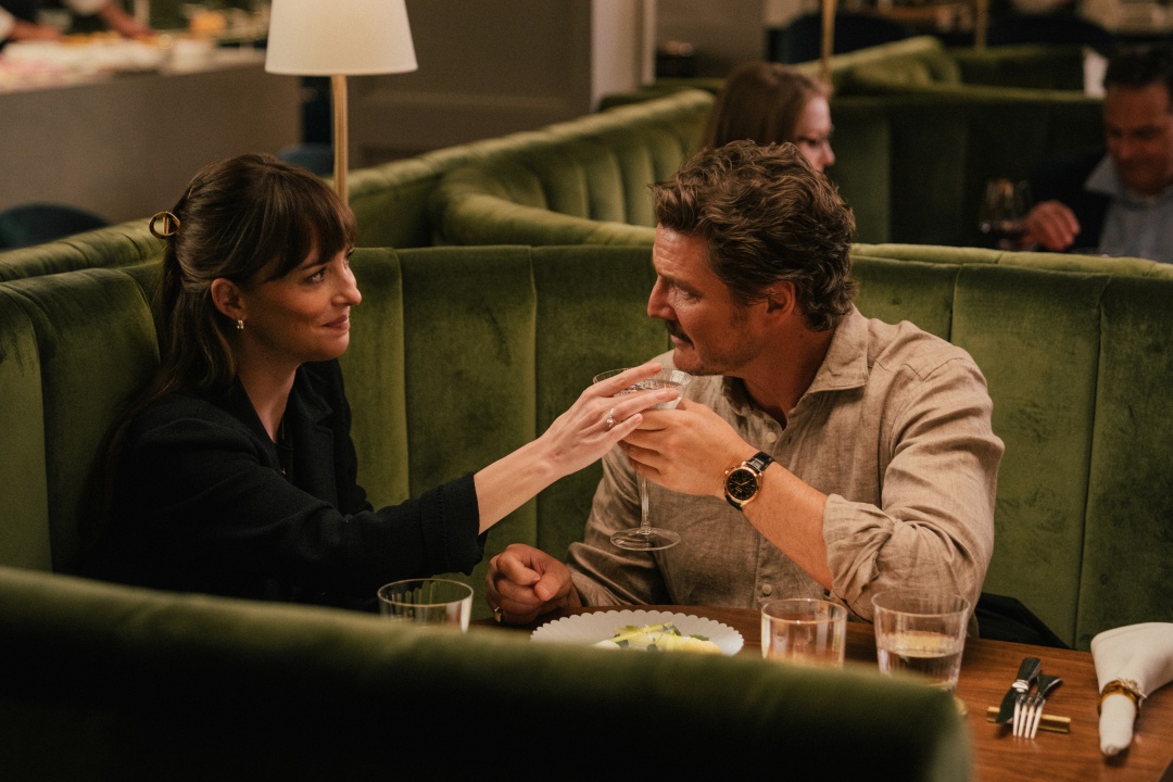 A woman and a man sit across from each other at a restaurant table, clinking glasses and smiling amid green velvet booths.