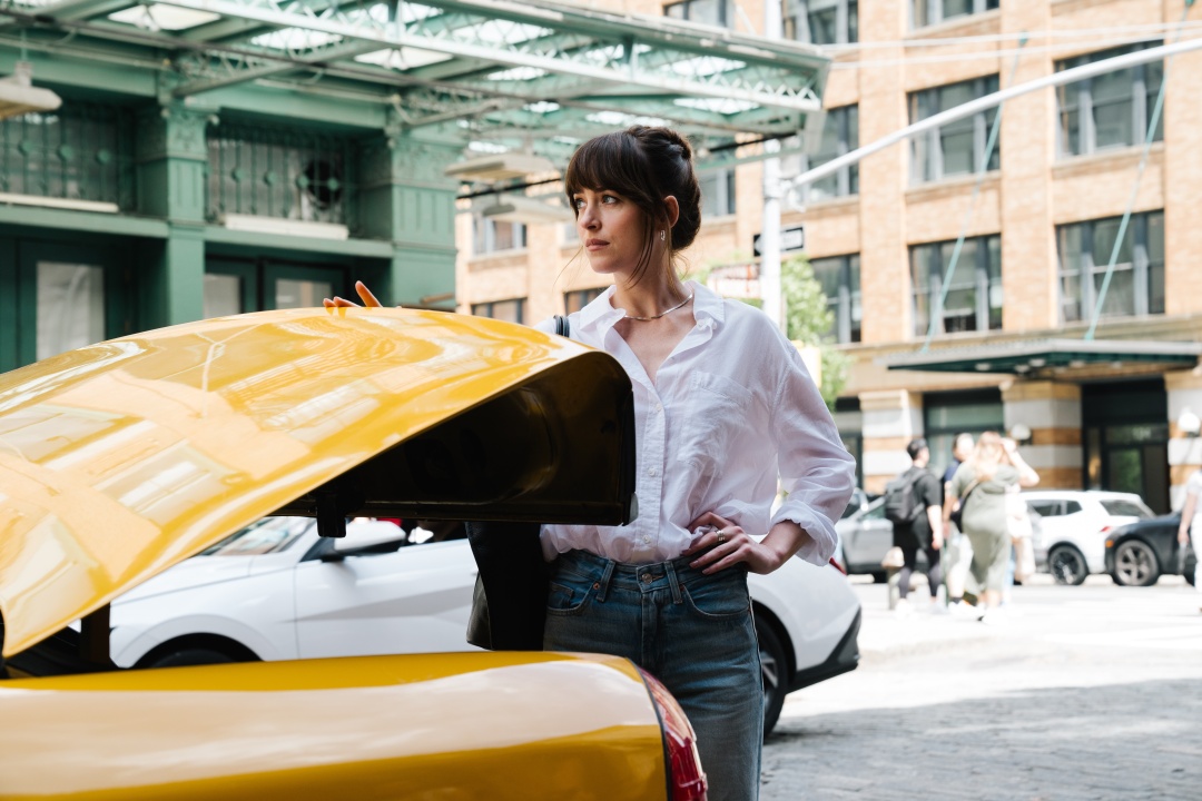A woman in a white shirt and jeans stands by an open yellow car trunk on a city street with buildings and people in the background.