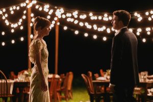 Two people stand facing each other outdoors at night, with string lights overhead and tables and chairs set up for an event in the background.