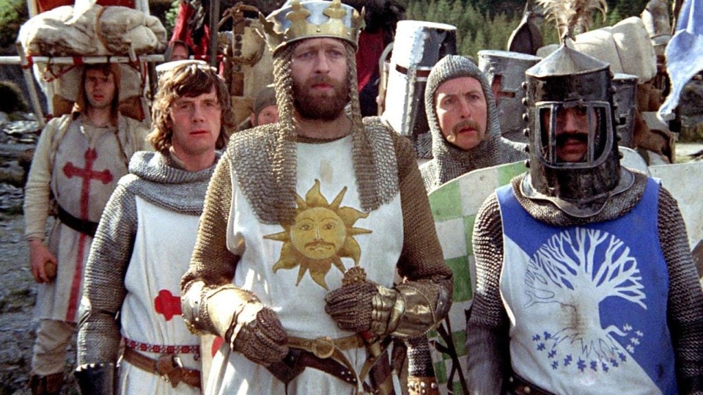 A group of men in medieval armor and costumes stand together outdoors, with one wearing a crown and a tunic featuring a sun emblem.