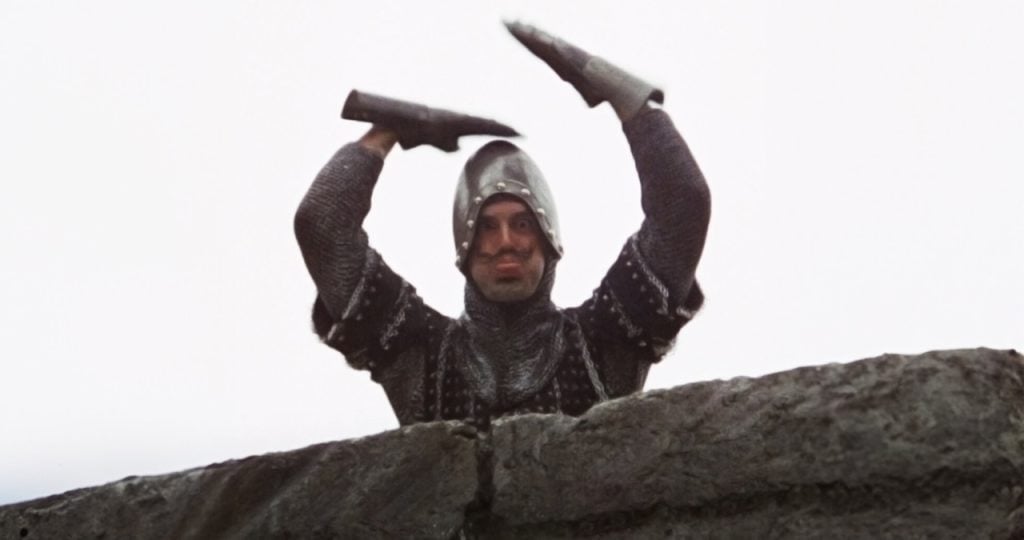 A person in medieval armor stands behind a stone wall, raising both arms with bent elbows against a white sky.