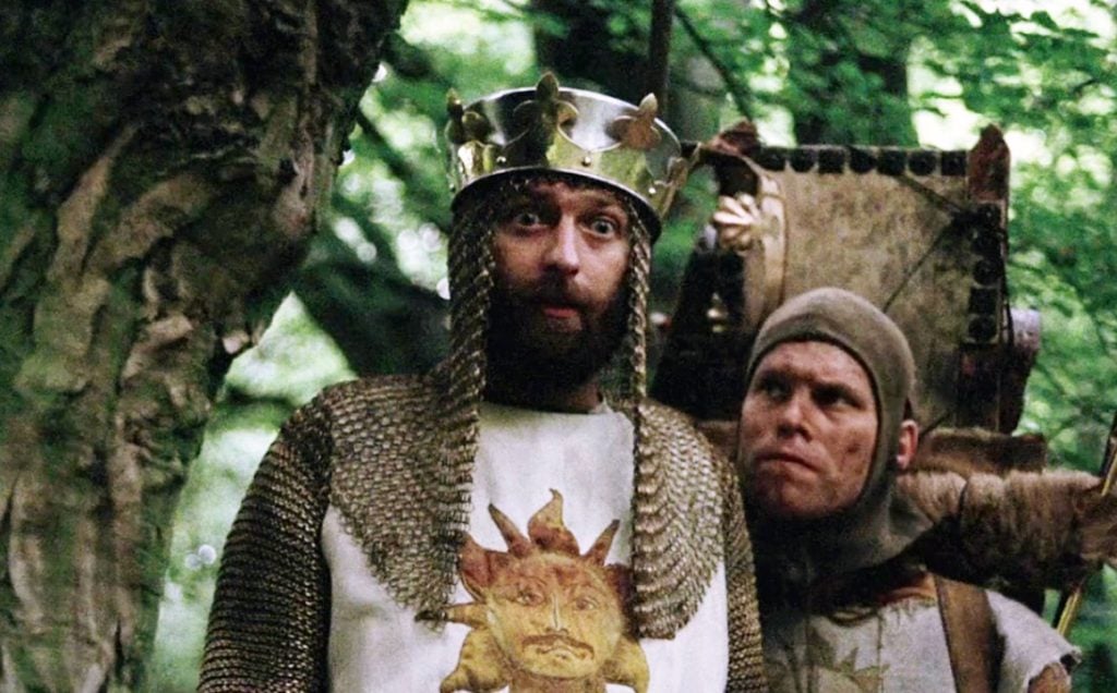 Two men in medieval costumes stand in a forest; one wears a crown and chainmail with a sun emblem, the other is in armor and carries a large pack.