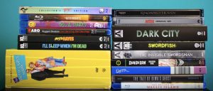 No Streaming Required: Physical Media Spotlight For The Week Of June 3rd A collection of Blu-ray and DVD cases are stacked and lined up, showing various movie and TV series titles, including "Kingdom of Heaven" and "Swordfish," against a blue background.