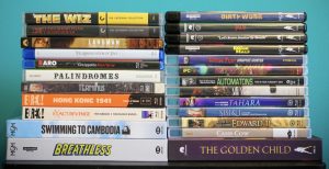 No Streaming Required: Physical Media Spotlight For The Week Of June 17th – Vinegar Syndrome, Criterion & More A collection of various DVD and Blu-ray movie cases stacked on a table against a blue background.