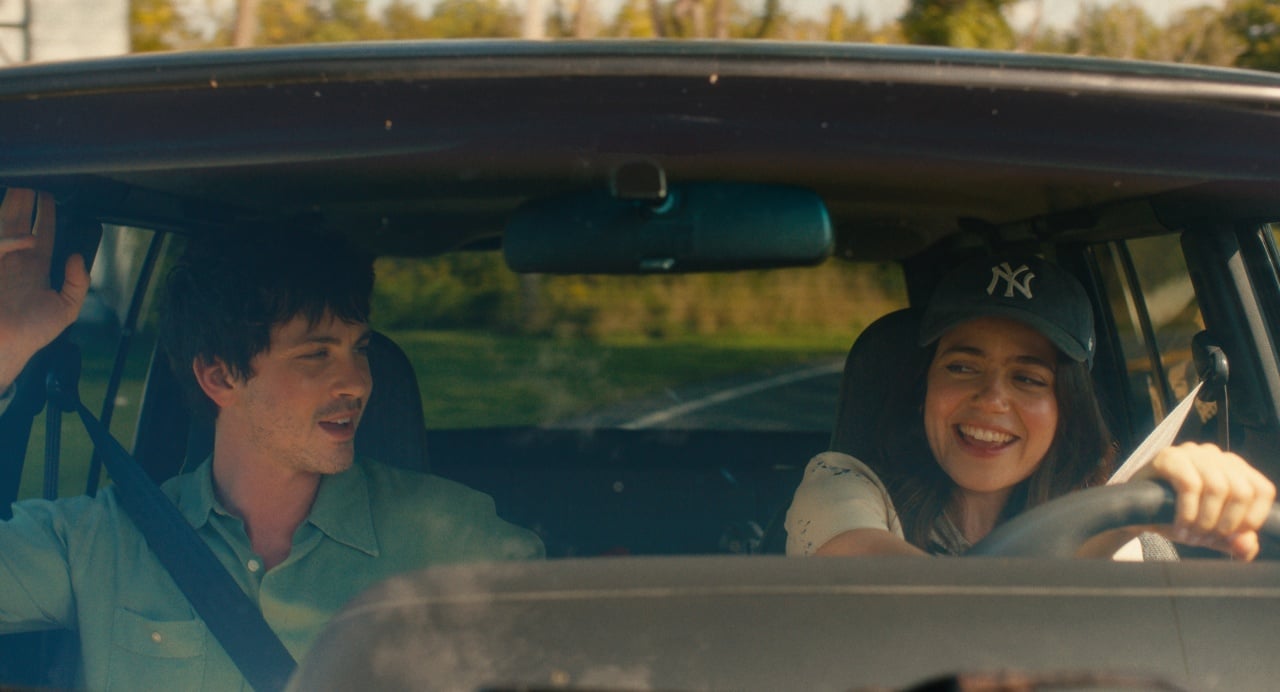 A young man and woman are sitting in the front seats of a car, with the woman driving and both appearing to enjoy a conversation while the car travels down a road.