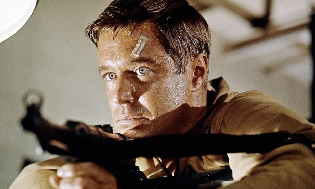 A man with a bandage on his forehead aims a rifle, looking intensely forward in a close-up shot.