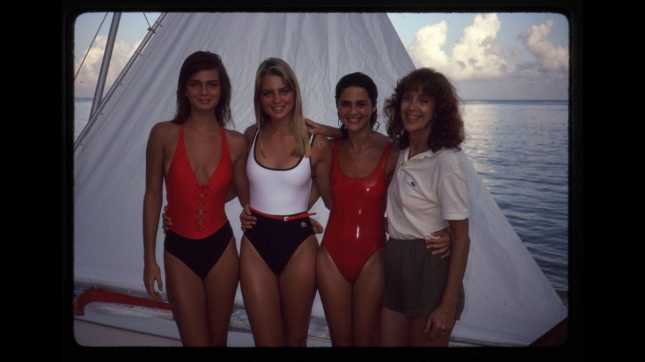 Four women pose and smile in front of a sailboat by the water, three wearing swimsuits and one wearing a white polo shirt and shorts.