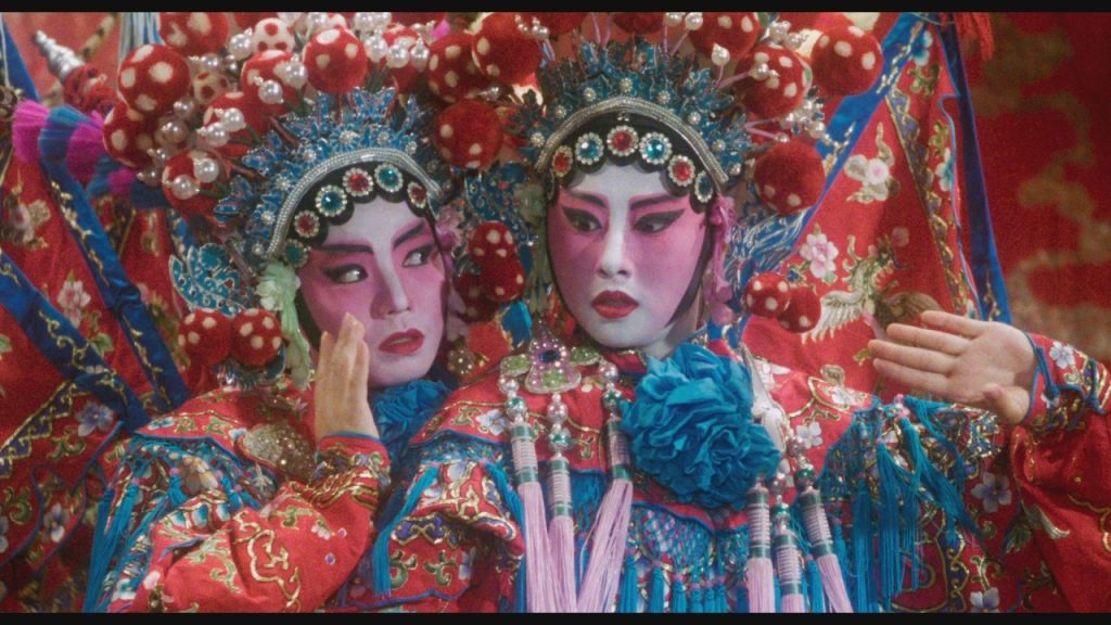 Two performers in ornate, colorful Chinese opera costumes and elaborate headdresses pose with dramatic facial expressions and makeup.