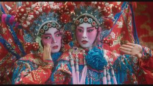Two performers in ornate, colorful Chinese opera costumes and elaborate headdresses pose with dramatic facial expressions and makeup.