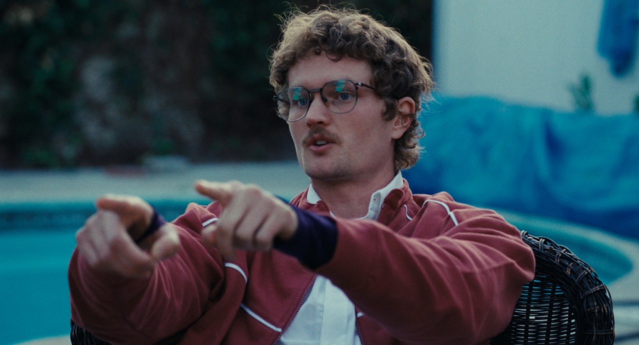 A man with curly hair and glasses sits in a wicker chair by a pool, wearing a red jacket and pointing with both hands.