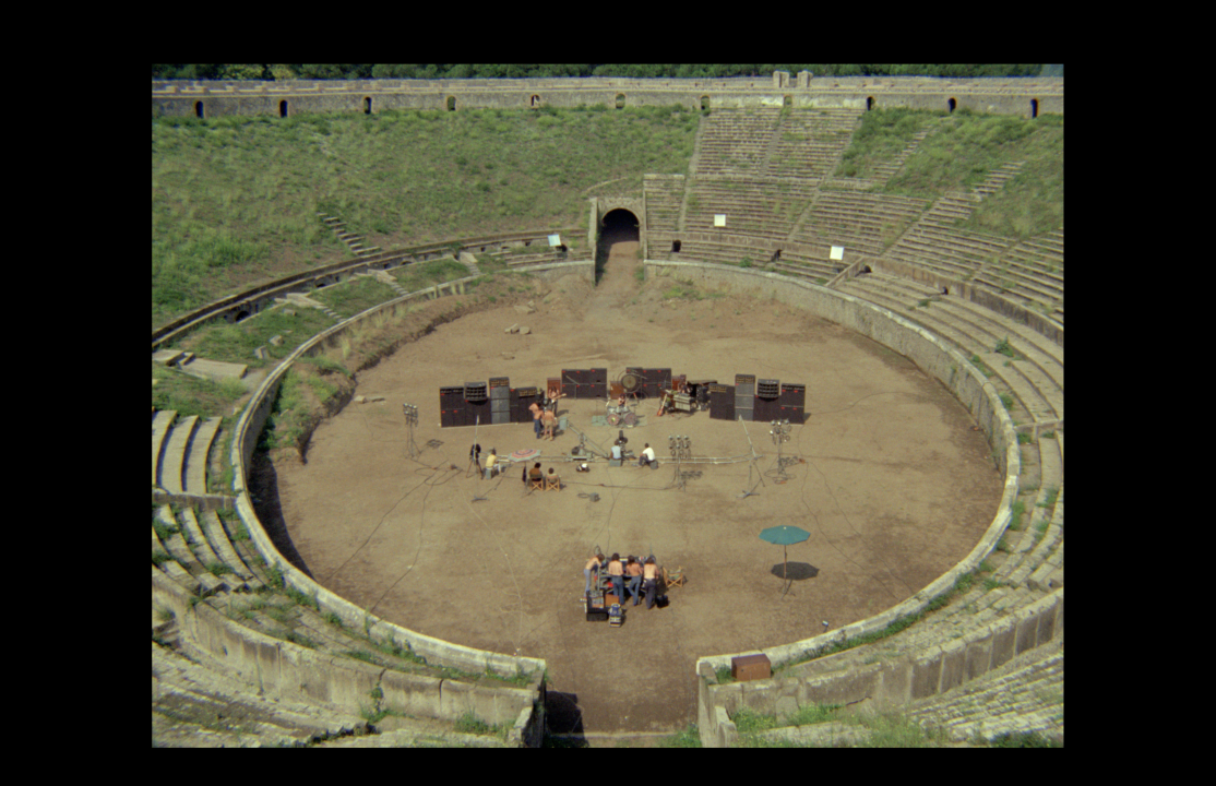 A small group of people sets up musical equipment in the center of an empty, ancient amphitheater with weathered stone seating and grass on the upper levels.