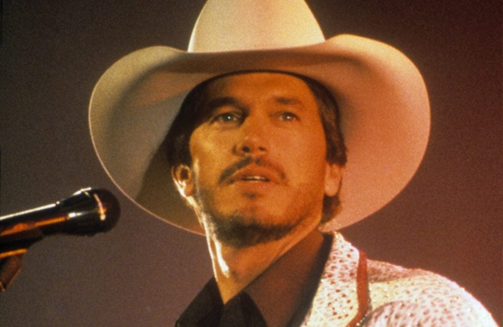 A man in a white cowboy hat and patterned jacket stands in front of a microphone, looking slightly upward against a reddish-brown background.