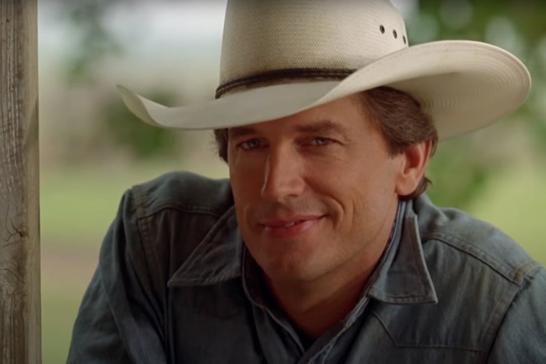 A man wearing a white cowboy hat and denim shirt smiles slightly while sitting outdoors near a wooden post.