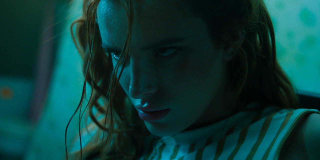 A person with long reddish hair and a serious expression looks intensely forward under blue-green lighting.