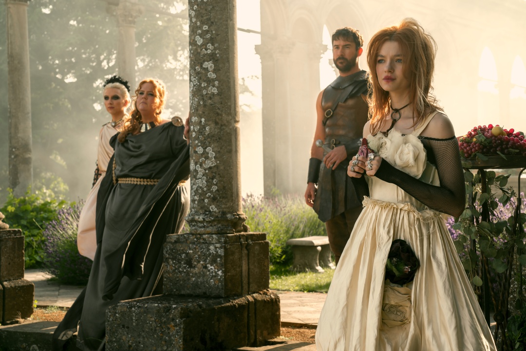 Four people in elaborate, ancient-style costumes stand among stone columns and greenery, with sunlight streaming through an old, open-air structure.