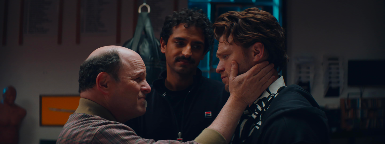 A bald man gently holds another man's face while a third man stands behind them, observing, in a dimly lit indoor setting.