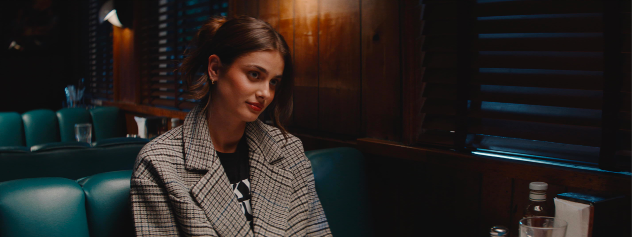 A woman with brown hair in a plaid coat sits in a dimly lit diner booth, looking to the side with a slight smile.