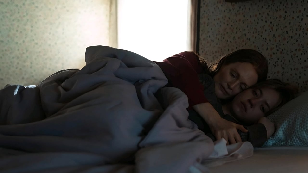 Two people lie in bed under a blanket, with one person hugging the other from behind. The room is dimly lit by a window in the background.