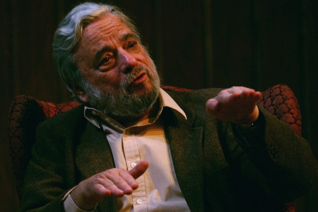 An older man with gray hair and a beard sits in a patterned chair, gesturing with both hands while speaking, against a dark, wooden background.