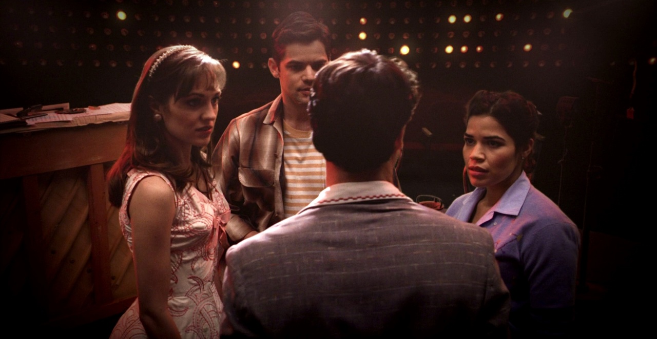 Four people stand closely together in conversation in a dimly lit room, with a piano and lights visible in the background.