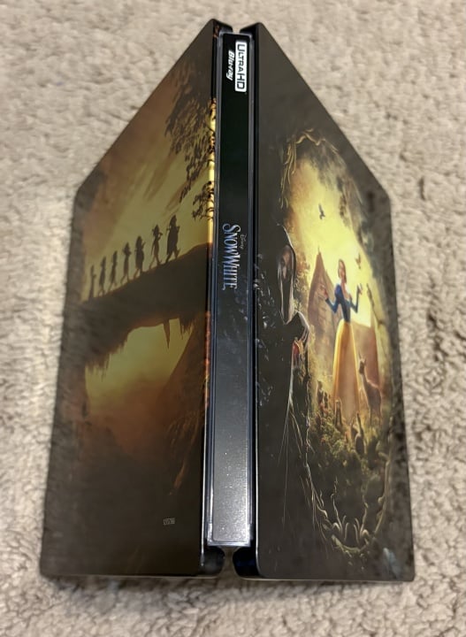 A Disney Snow White steelbook case stands upright on a beige carpet, showing the dwarfs on one side and Snow White with birds on the other.