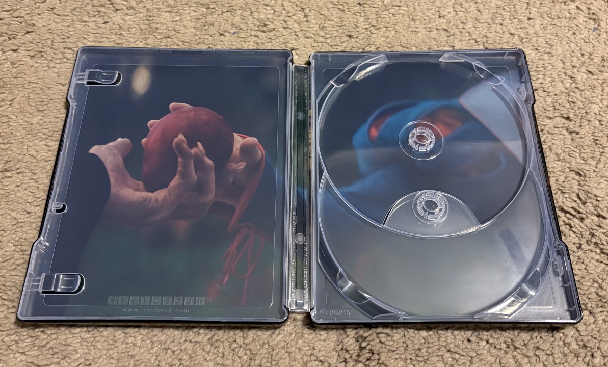 Open Blu-ray steelbook case on a beige carpet, showing a hand holding a red apple on the left and an empty disc holder on the right.