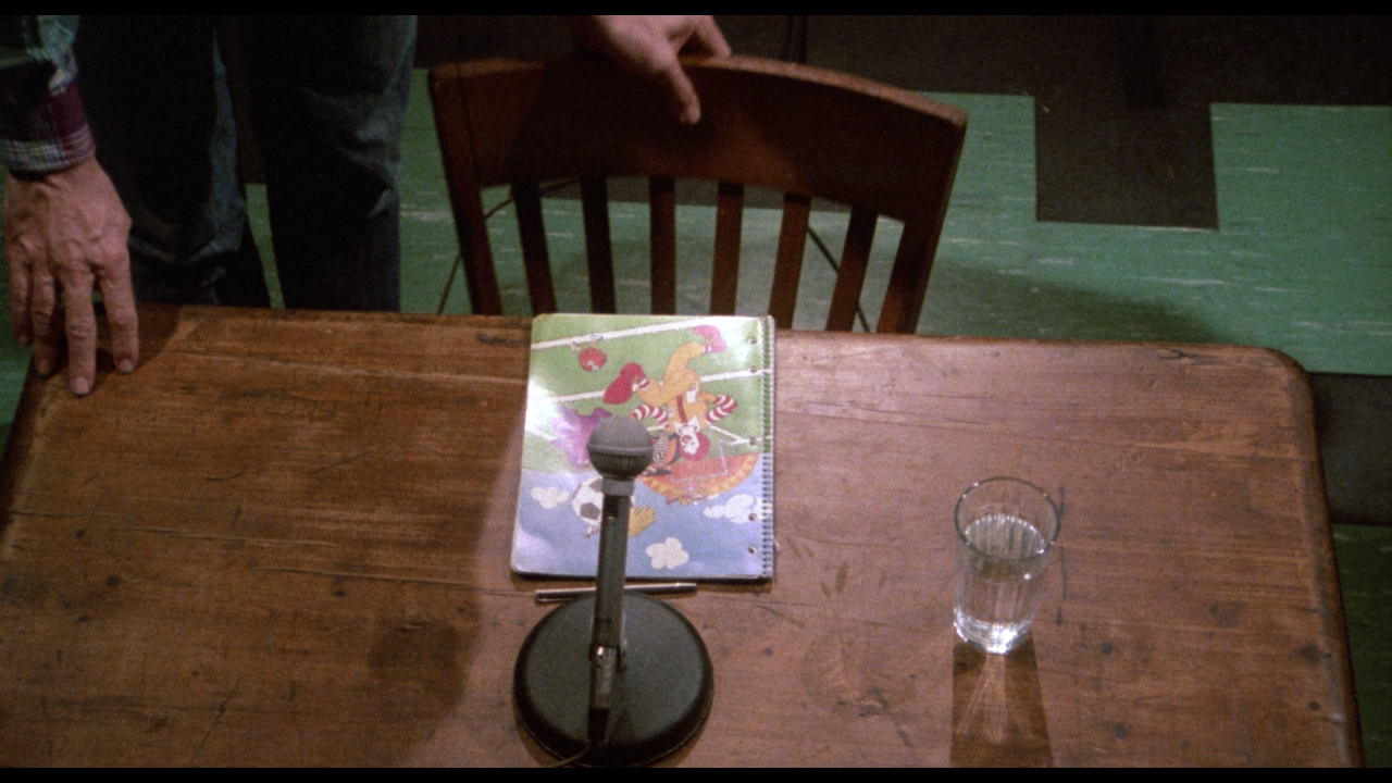 A hand pulls out a wooden chair at a table with a microphone, a glass of water, and a colorful illustrated notebook.