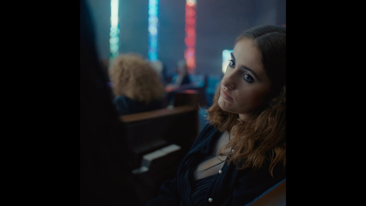 A young woman with wavy brown hair and dark eye makeup sits in a pew, looking slightly to the side in a dimly lit room with blurred colorful lights in the background.