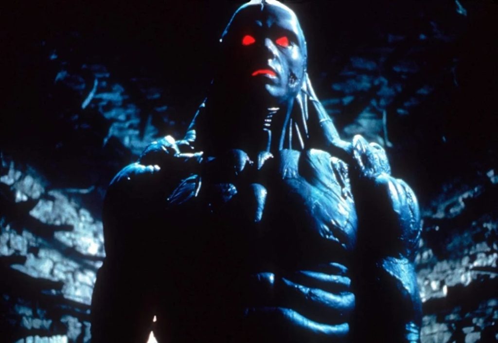 A humanoid figure with glowing red eyes and a muscular, armored body stands in a dim, blue-lit environment.