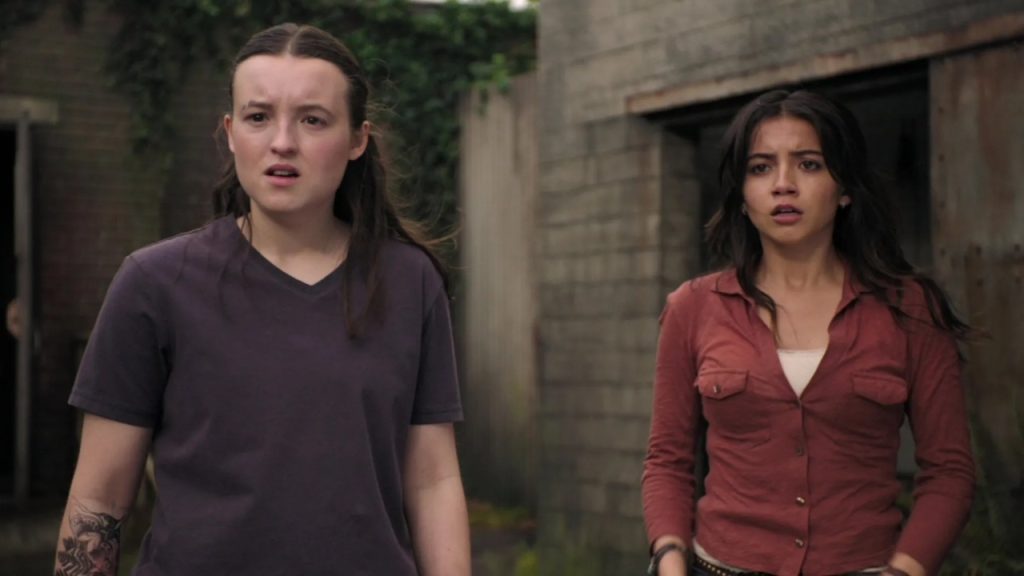 Two young women stand outside, looking ahead with concerned expressions, in front of a concrete building with overgrown vegetation.