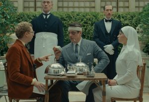 A man with bandages on his face sits at a table with food, accompanied by a nurse, another man, and two butlers standing behind them, in an outdoor garden setting.