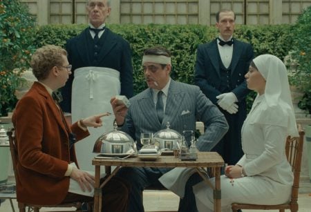 A man with bandages on his face sits at a table with food, accompanied by a nurse, another man, and two butlers standing behind them, in an outdoor garden setting.