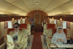 A man in sunglasses reads a newspaper and a woman in white sits with a notebook on a retro-styled airplane; a flight attendant stands in the background.