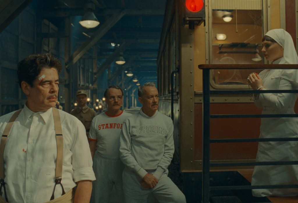 Several men in casual and athletic clothing stand near a vintage train car while a woman dressed as a nun stands on the train steps, holding her hands together.