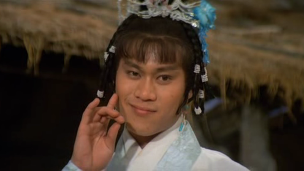 Person wearing traditional Chinese attire and headdress, smiling and posing with one hand near their face, in front of a thatched-roof background.