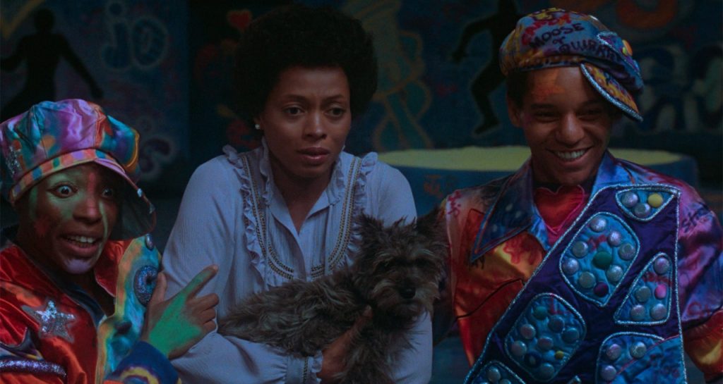 Three people stand together, one holding a small dog. Two are wearing colorful, patterned outfits and hats, while the third wears a white top. A brightly painted mural is visible in the background.