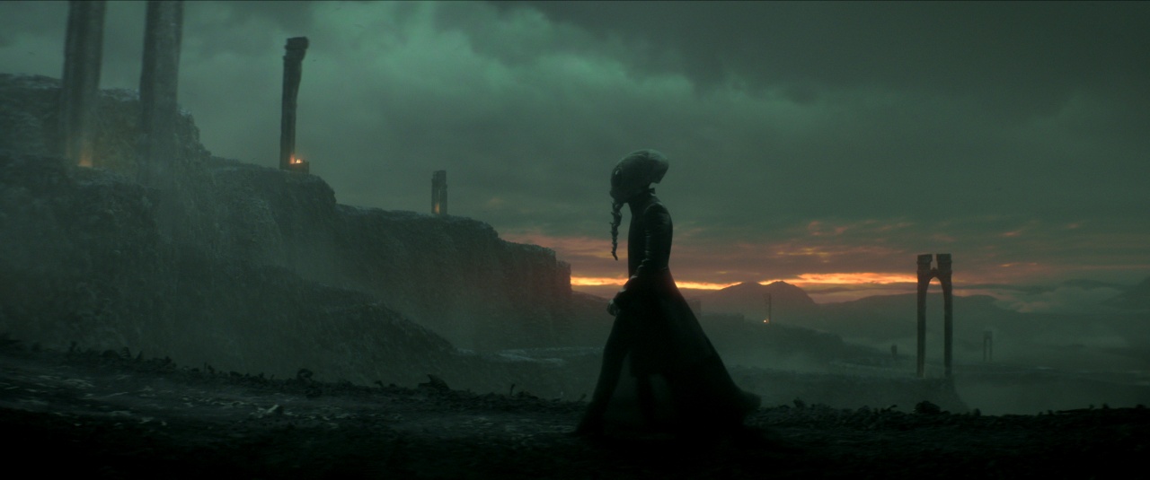 A person in a long cloak and headpiece walks along a dark, misty ruin at sunset, with stone pillars and mountains in the background.